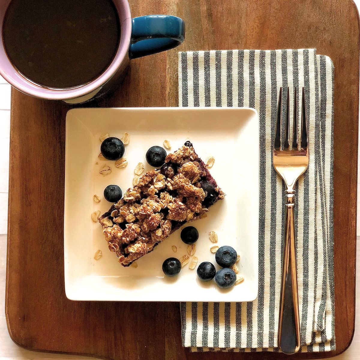 Blueberry oatmeal bars on a white plate with a cup of coffee, napkin, and fork nearby. 