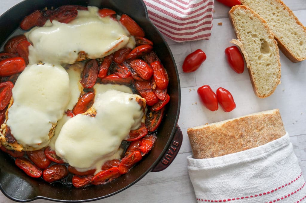 chicken breast in skillet covered with melted Mozzarella cheese and tomatoes. Loaf of crusty bread has been sliced to the side.