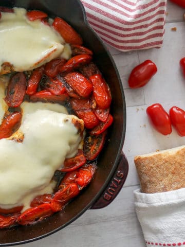 chicken breast in skillet covered with melted Mozzarella cheese and tomatoes. Loaf of crusty bread has been sliced to the side.