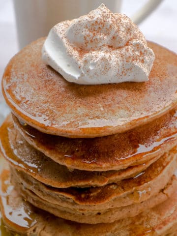 Tall stack of gingerbread pancakes topped with maple syrup, whipped cream, and gingerbread spices.