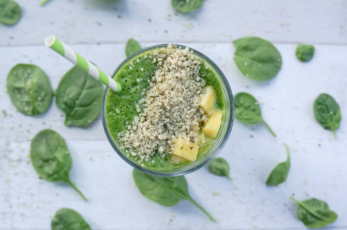 Bright green smoothie in a tall glass topped with pineapple and hemp seeds. A cute green and white stripped paper straw is added for drinking.