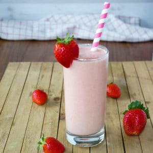 Pink smoothie in a tall glass with a strawberry garnished on the side. A pink and white stripped straw is in the glass. Apples and strawberries are seen in the background.