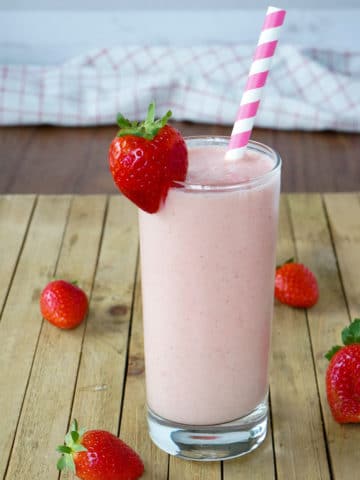 Pink smoothie in a tall glass with a strawberry garnished on the side. A pink and white stripped straw is in the glass. Apples and strawberries are seen in the background.