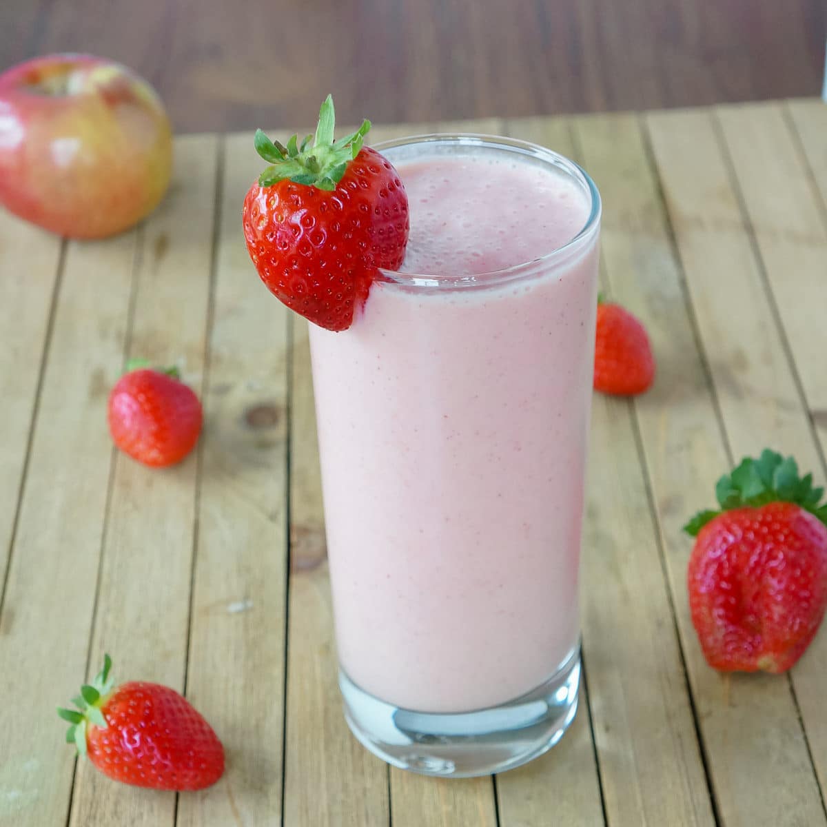 Pink smoothie in a tall glass with a strawberry garnished on the side. A pink and white stripped straw is in the glass. Apples and strawberries are seen in the background.