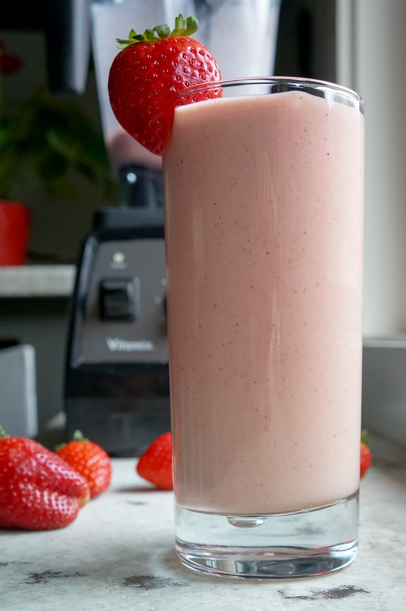 Pink smoothie in a tall glass with a strawberry garnished on the side. A pink and white stripped straw is in the glass. Apples and strawberries are seen in the background.