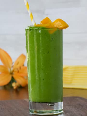 green spinach and mango smoothie in a tall glass with mango chunks on top and a yellow and white stripped straw.