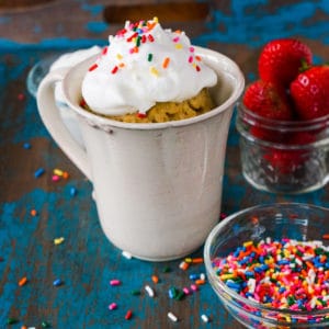 mug cake baked in a coffee mug with whipped cream and sprinkles on top. sprinkles and strawberries are in the background.