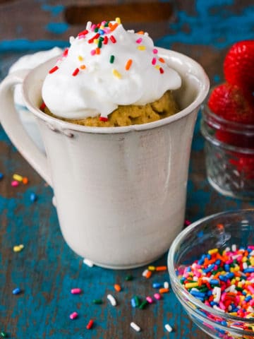 mug cake baked in a coffee mug with whipped cream and sprinkles on top. sprinkles and strawberries are in the background.