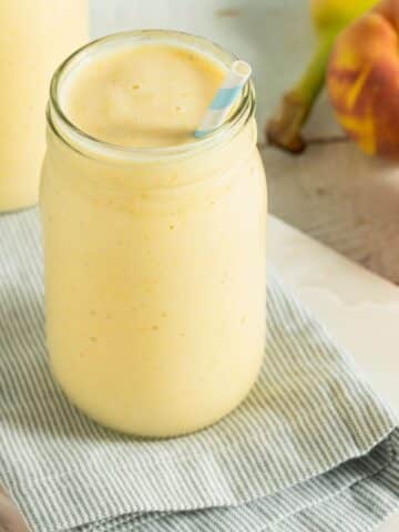 A cold and creamy pineapple peach smoothie in a clear glass cup with a blue and white striped straw.