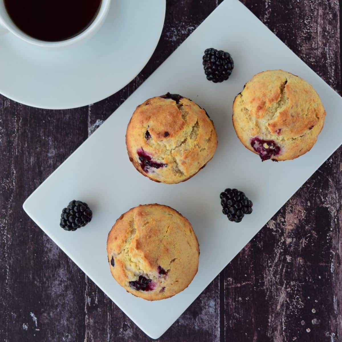 3 homemade blackberry muffins on a white plate with fresh blackberries and a cup of coffee in the background